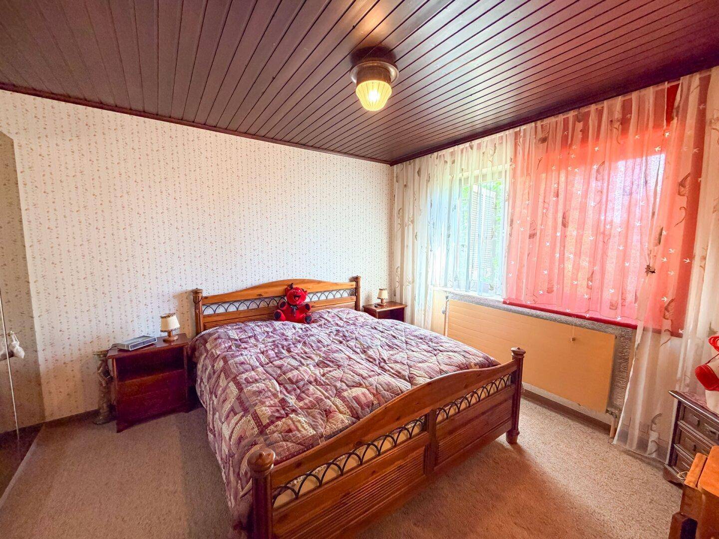 Schlafzimmer mit einem Doppelbett aus Holz, gemusterter Tapete und einem Fenster mit roten Vorhängen.