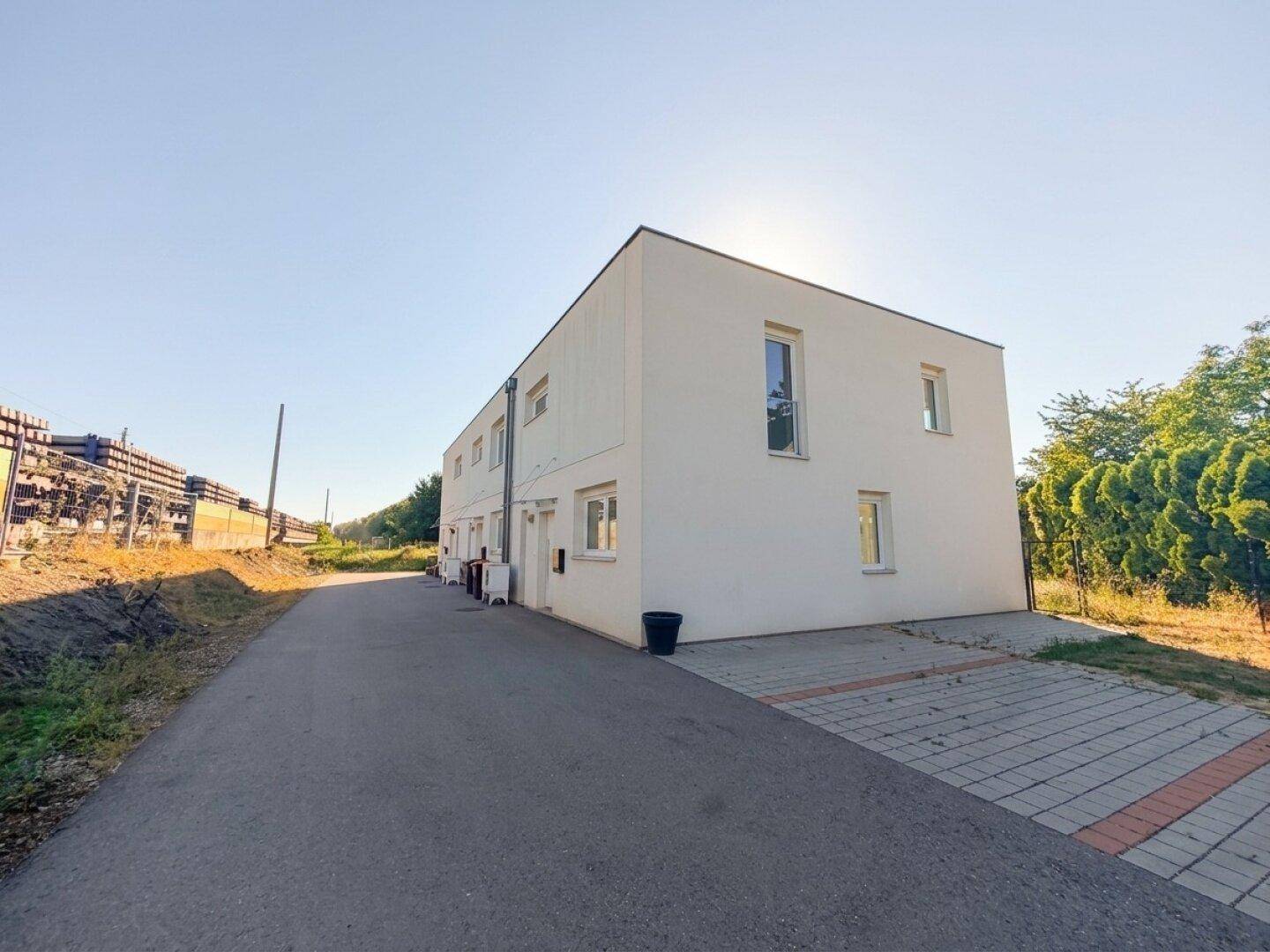 Modernes Gebäude mit weißer Fassade, Flachdach und gepflastertem Parkplatz unter blauem Himmel.