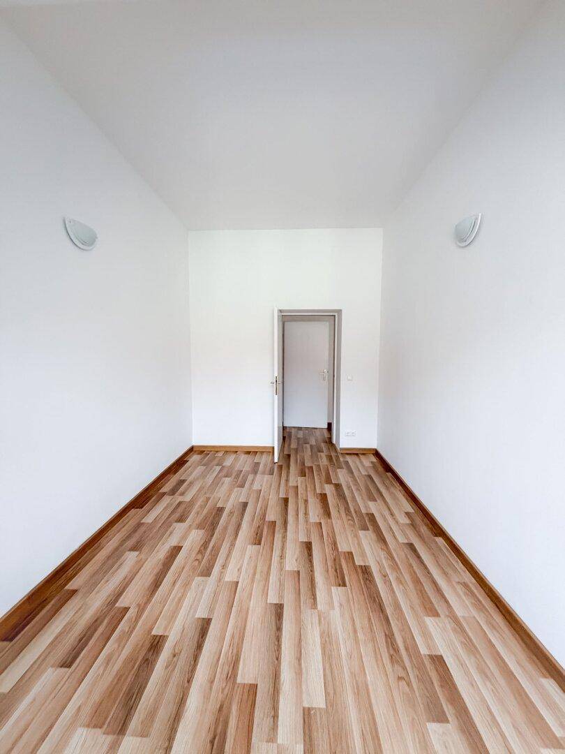 Leeres Zimmer mit Laminatboden und weißen Wänden, das durch eine offene Tür zugänglich ist.