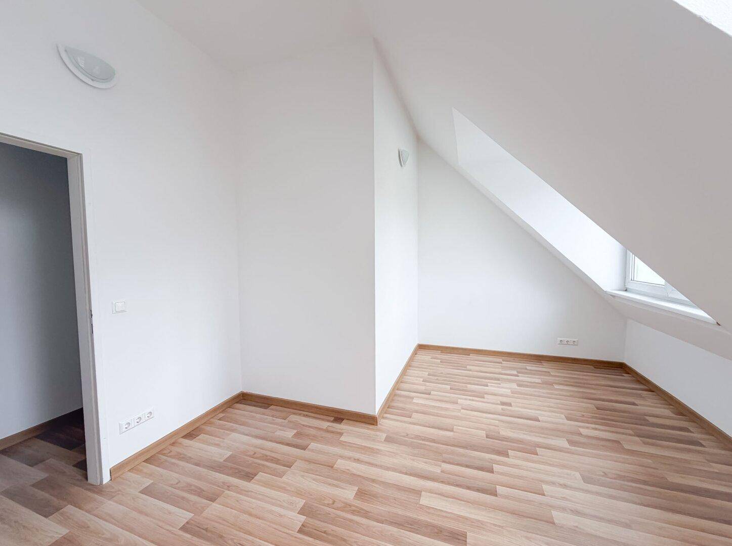 Leeres Zimmer mit Laminatboden und weißen Wänden, teilweise mit Dachschräge und Türzugang.