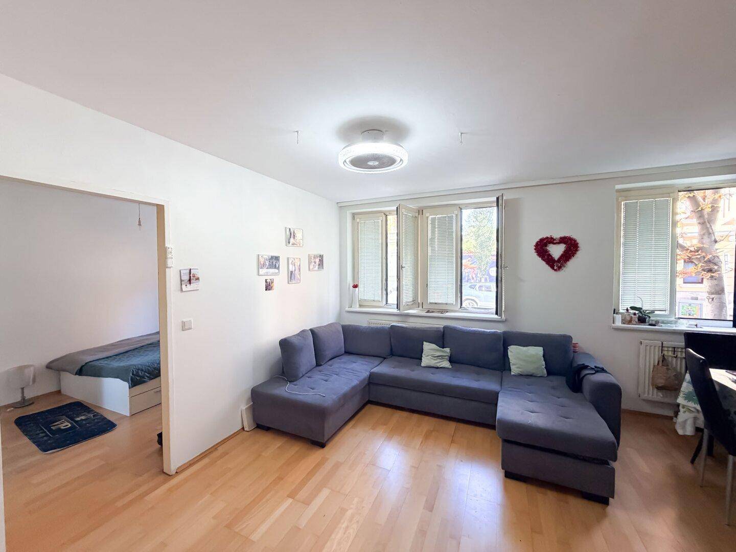 Wohnzimmer mit Ecksofa und Blick in ein angrenzendes Schlafzimmer, ausgestattet mit Holzboden.