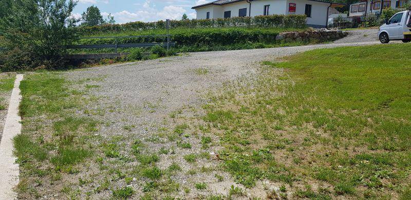 Weitläufige Schotterfläche mit Grasbewuchs, die zu einer Zufahrt und einem Haus im Hintergrund führt.
