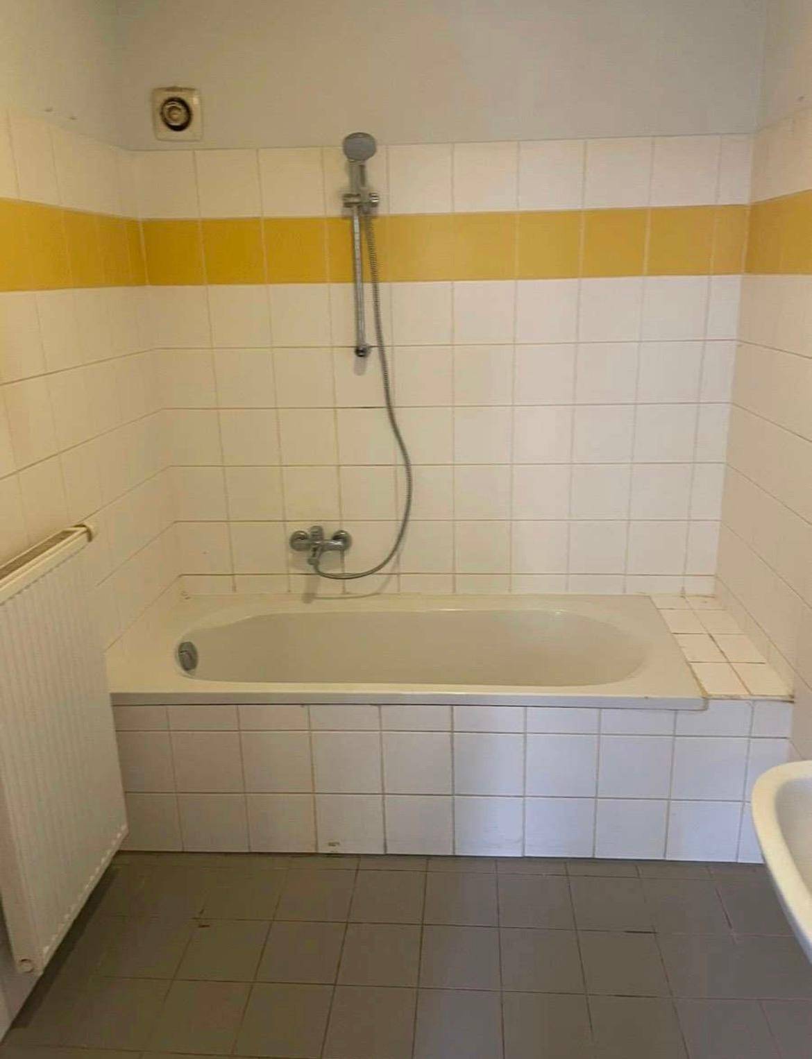 Badezimmer mit Badewanne, Duscharmatur und weißen Fliesen mit gelbem Akzentstreifen.