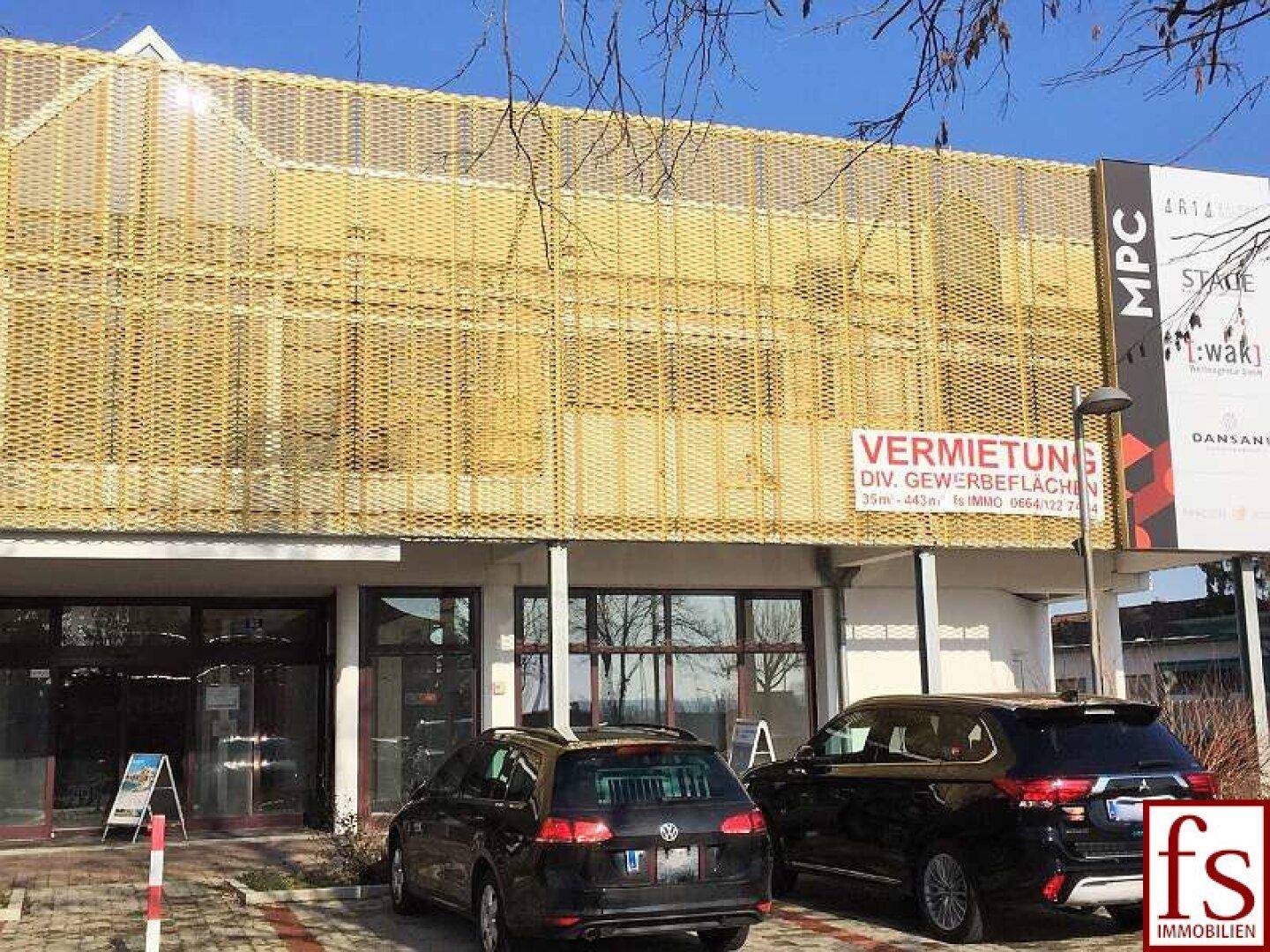 Seitenansicht des Gewerbegebäudes mit goldener Gitterfassade und Parkplätzen im Vordergrund unter klarem Himmel.