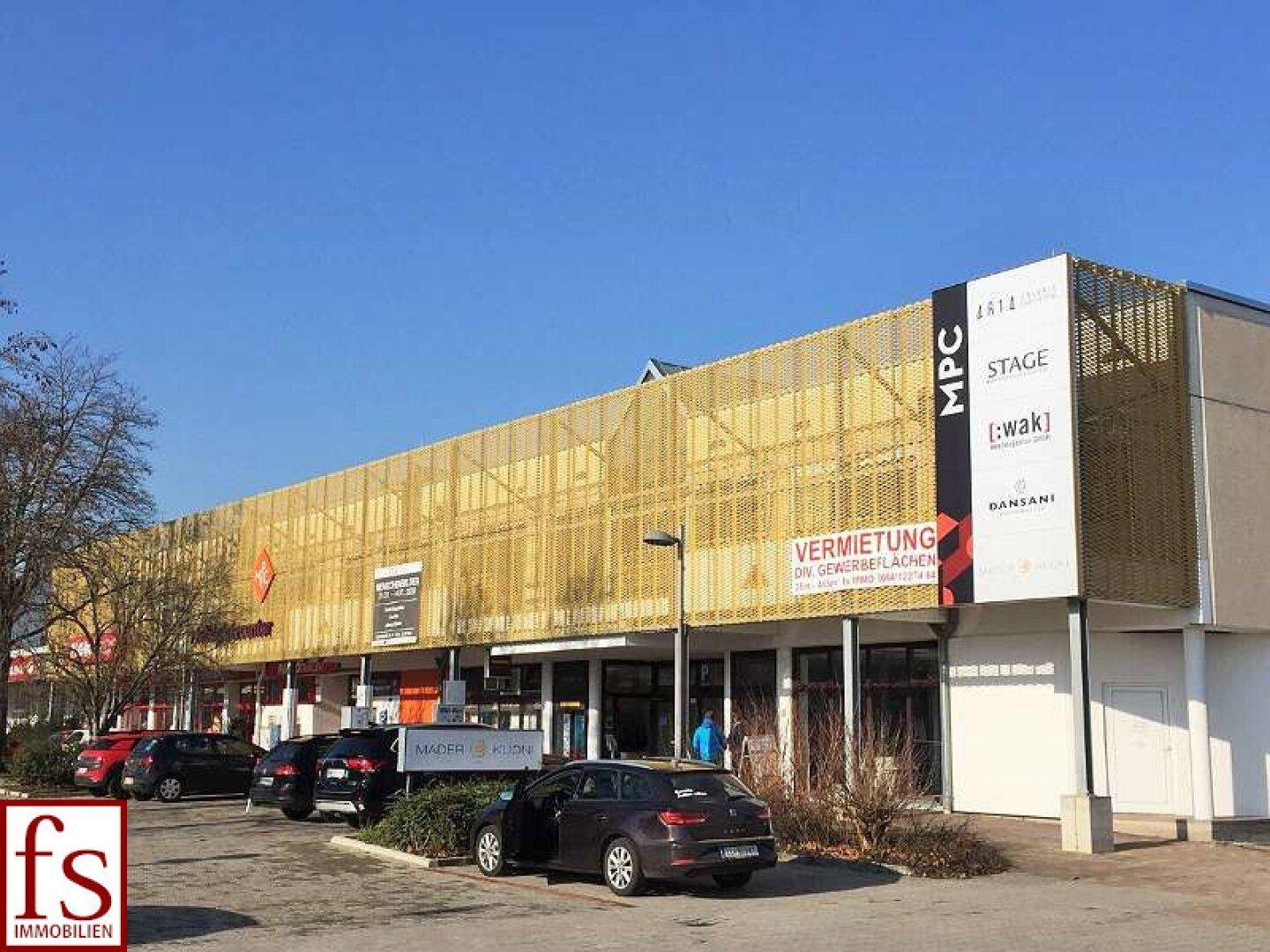Ansicht des Gewerbegebäudes mit goldener Fassade und Hinweisschildern zur Vermietung von Flächen und Parkplätzen.