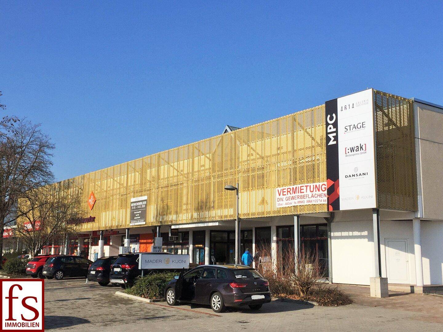 Ansicht des modernen Einkaufszentrums mit goldener Fassade, Geschäften und Parkplätzen im Vordergrund.