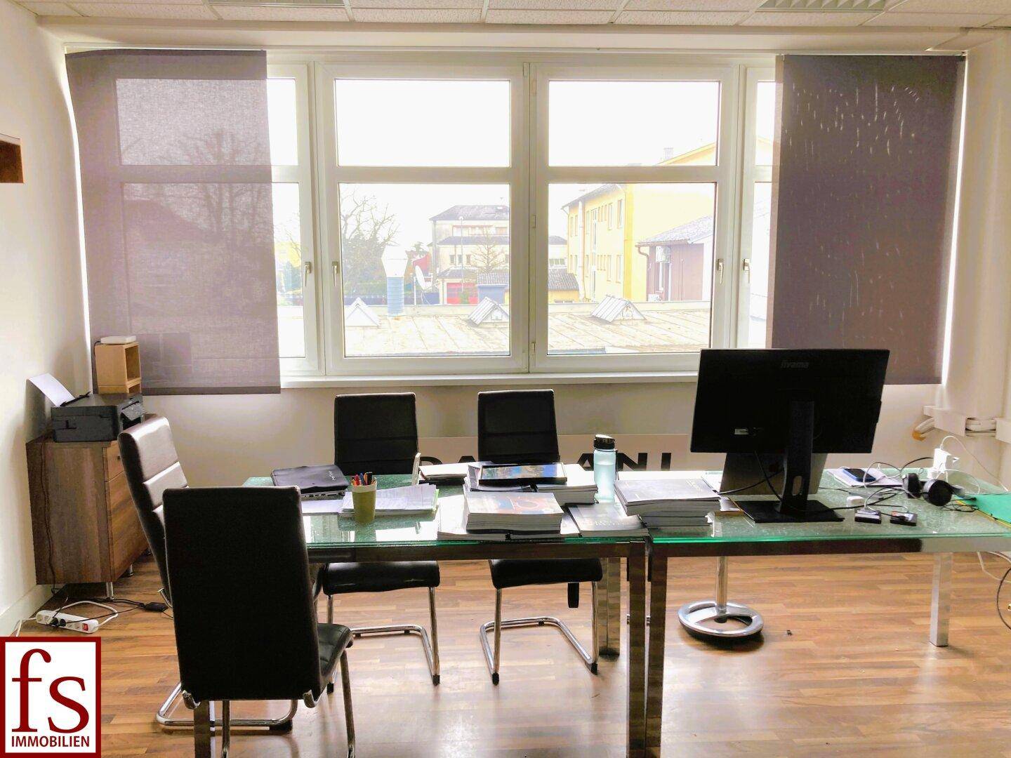 Helles Büro mit großem Fenster, Holzboden und modernem Glastisch, ideal für Besprechungen.