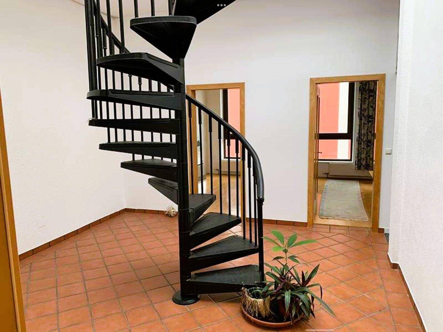 Eingangsbereich mit schwarzer Wendeltreppe, Terrakotta-Fliesenboden und Zimmerpflanze.