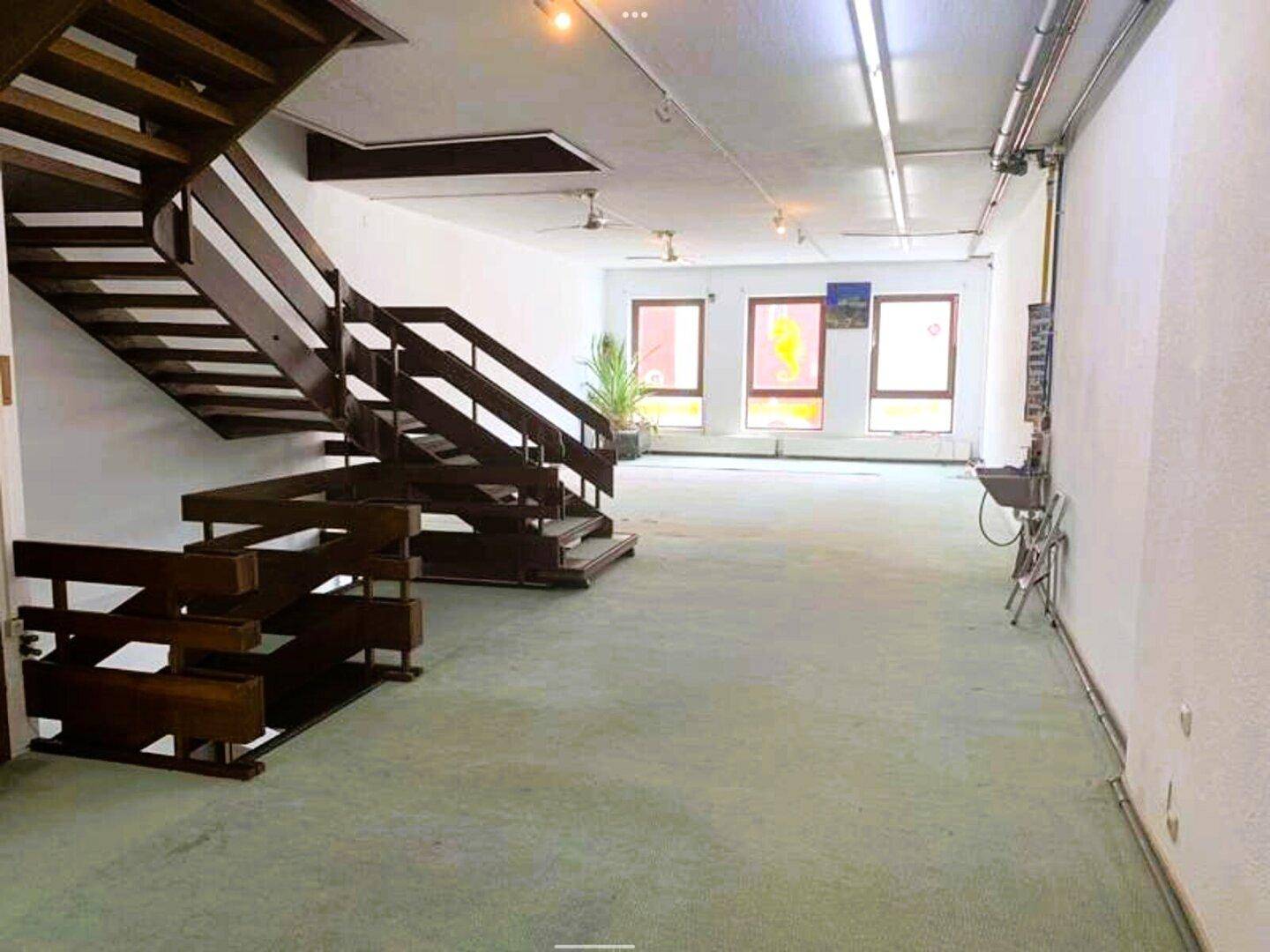 Heller Innenraum mit markanter Holztreppe, grünem Teppichboden und Fenstern im Hintergrund.