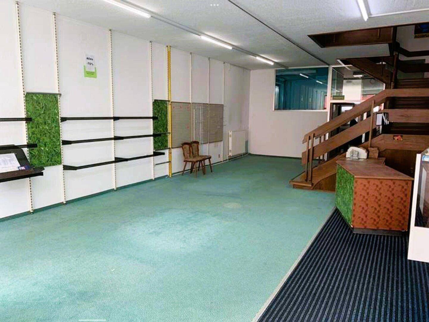 Geräumiger Innenraum eines Ladenlokals mit leeren Regalen, grünem Teppich und Holztreppe.