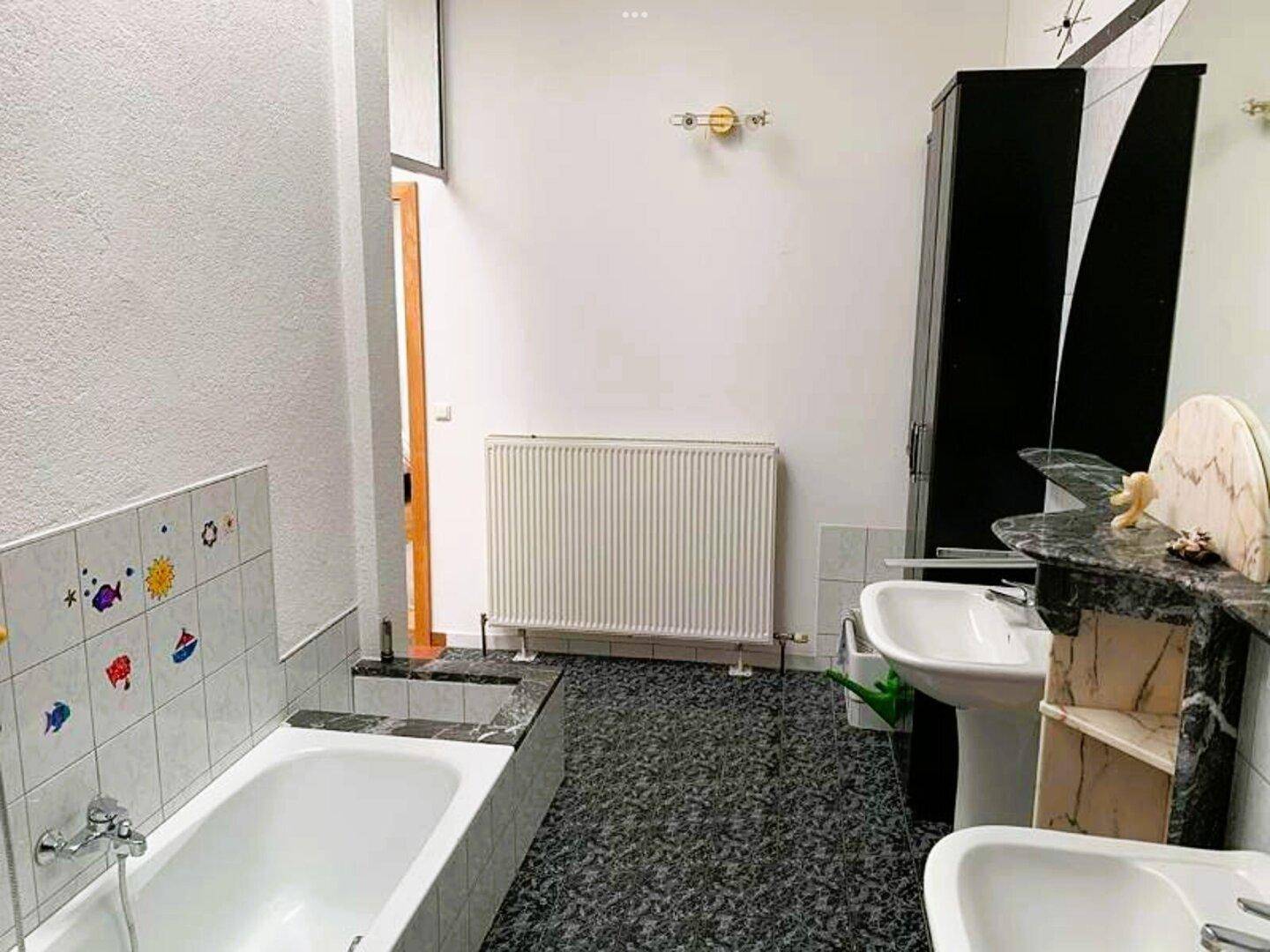 Badezimmer mit Badewanne, Doppelwaschbecken, Heizkörper und dunklem Fliesenboden.