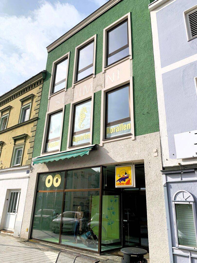 Grünes Gebäude mit großen Schaufenstern und Werbeschildern in einer städtischen Umgebung.
