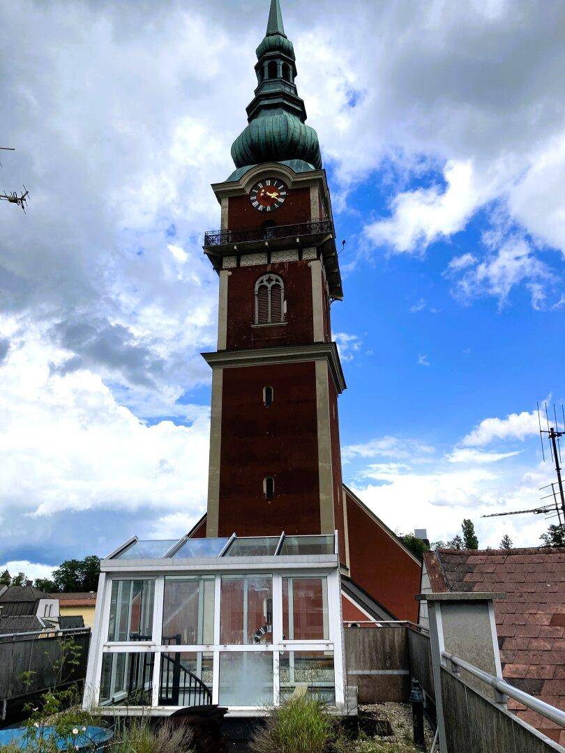 Hoher roter Kirchturm mit Uhr vor blauem Himmel und einem Wintergarten im Vordergrund.