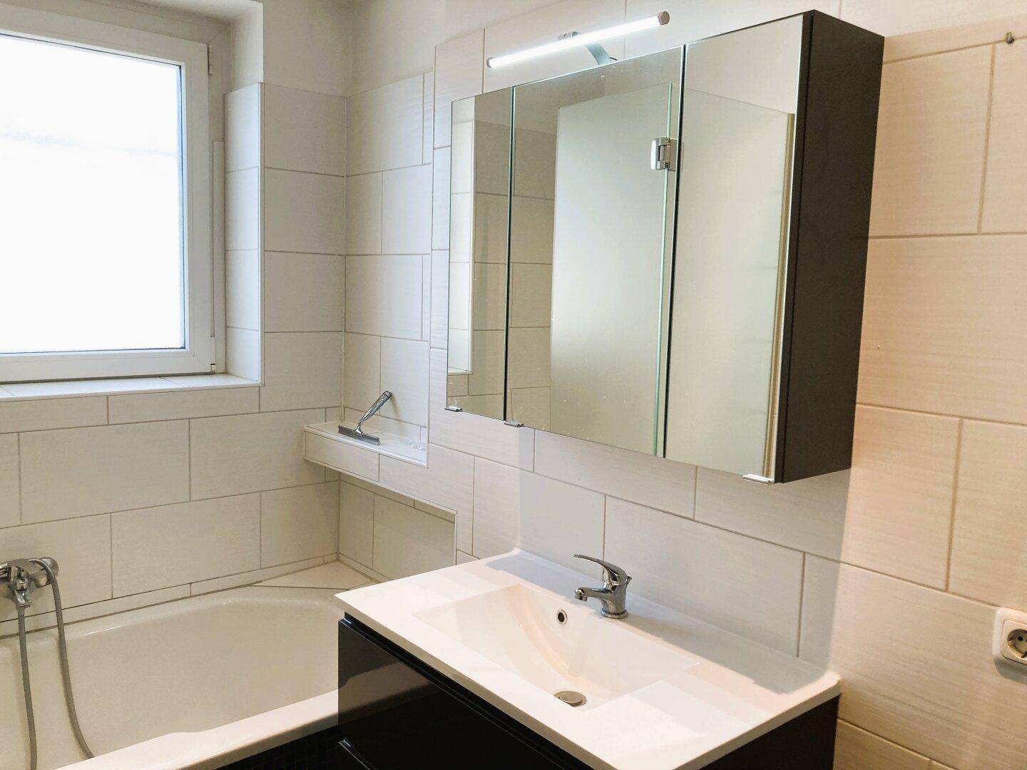 Helles Badezimmer mit Badewanne, Fenster, modernem Waschtisch und Spiegelschrank.