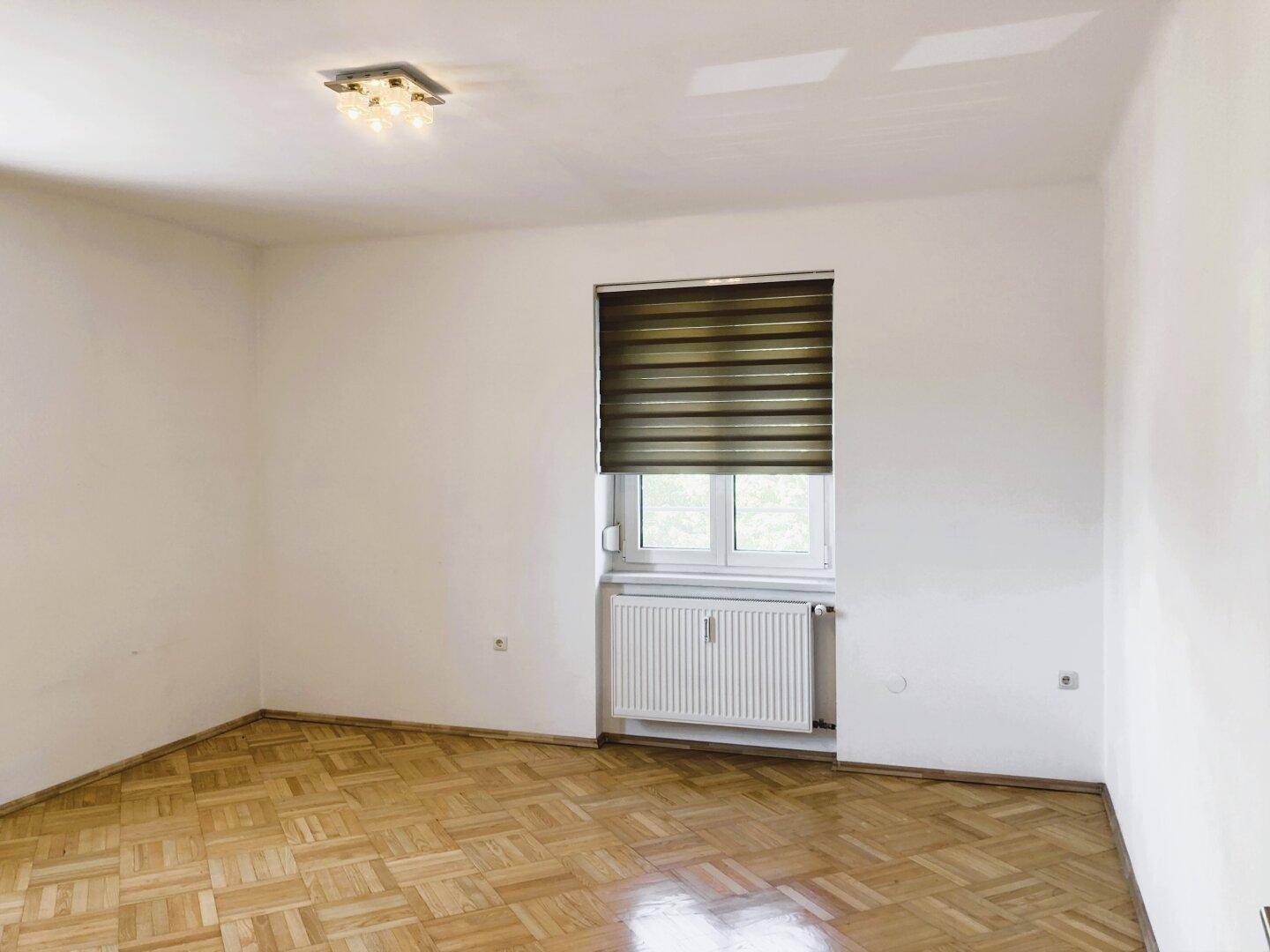 Helles Schlafzimmer mit Parkettboden, Heizkörper unter dem Fenster und neutralen Wänden.