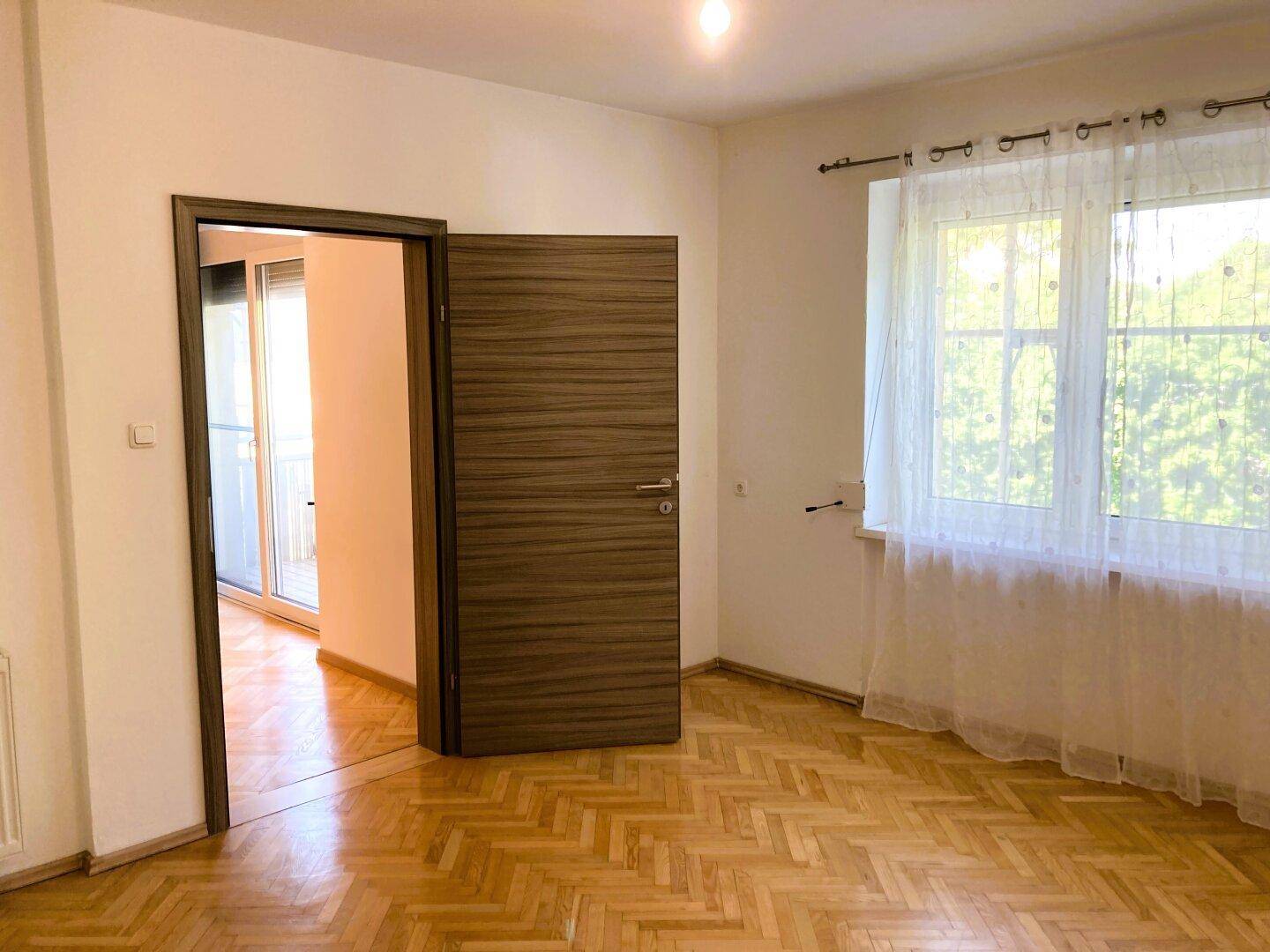 Schlafzimmer mit Parkettboden und großem Fenster, das viel Tageslicht hereinlässt.