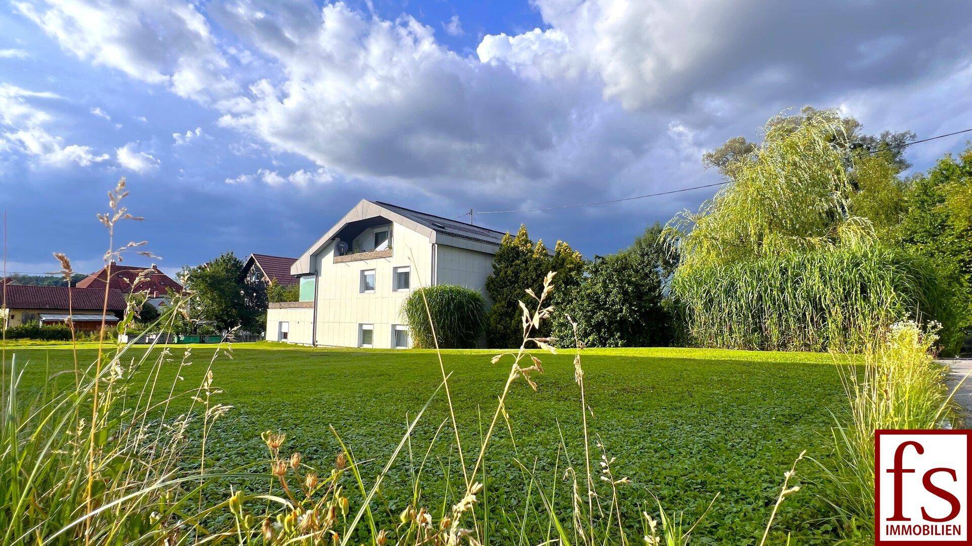 Hausansicht von der Ferne mit hohem Gras im Vordergrund und bewölktem Himmel.