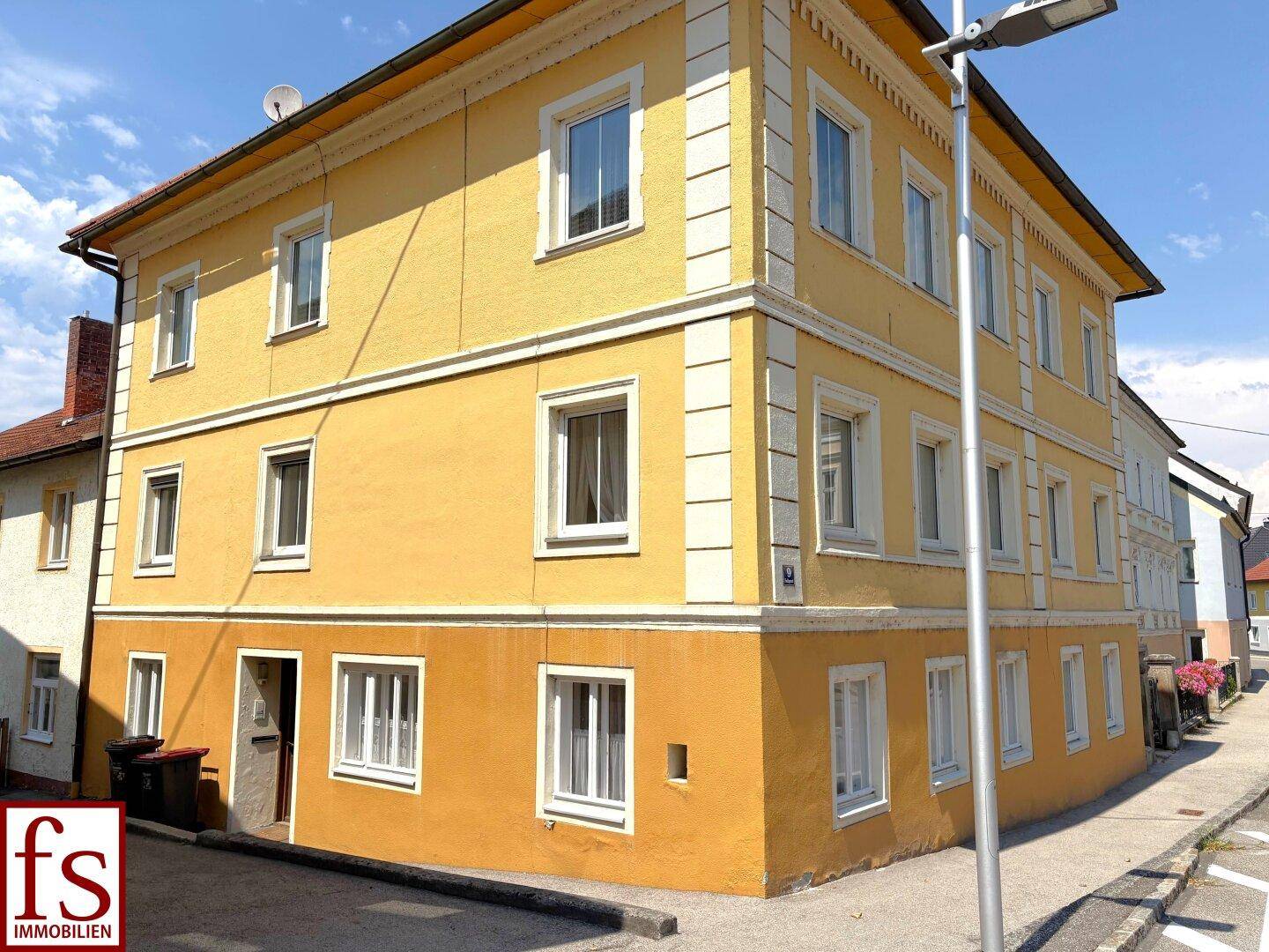 Ansicht des gelben Gebäudes von der Straßenseite mit Eingangsbereich und Mülltonnen.