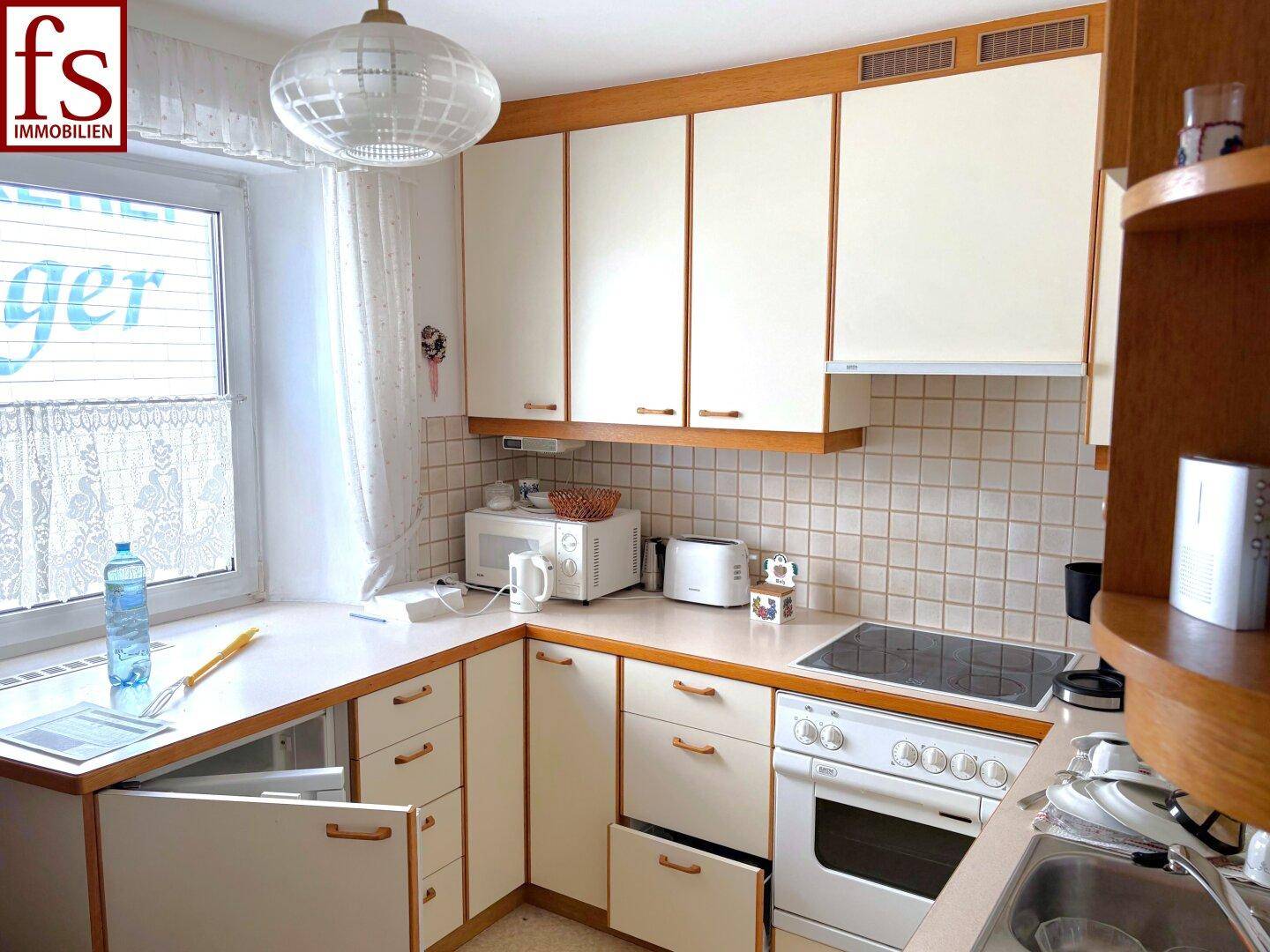 Kleine Küche mit hellen Holzschränken, Fenster und grundlegenden Küchengeräten.