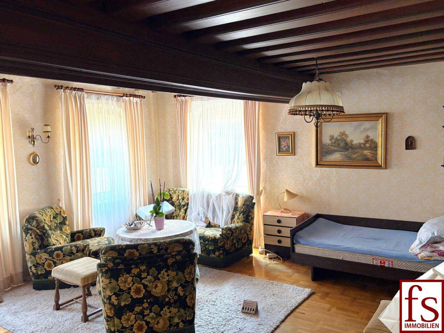 Gemütliches Wohnzimmer mit dunklen Deckenbalken, floralen Sesseln und einem Tagesbett.