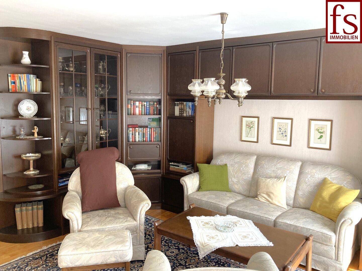 Wohnzimmer mit dunklen Einbauschränken, beigem Sofa und Sessel, dekoriert mit Bildern.