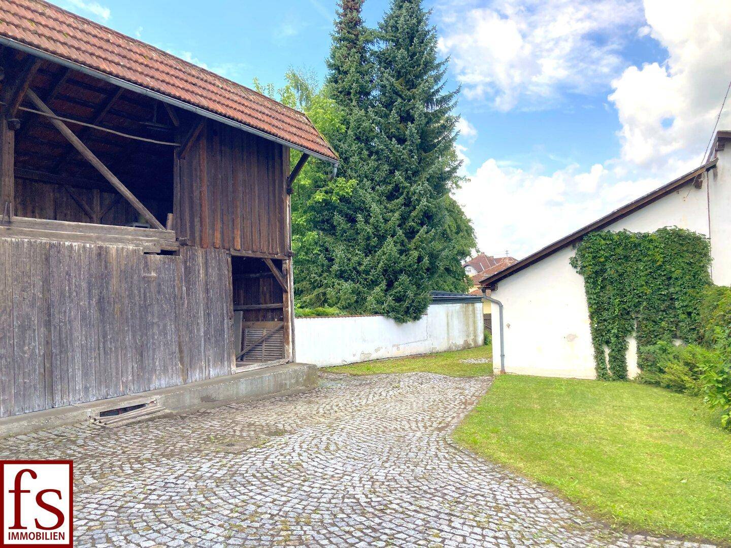 Gepflasterte Einfahrt neben einem Holzschuppen und einer Rasenfläche unter blauem Himmel.