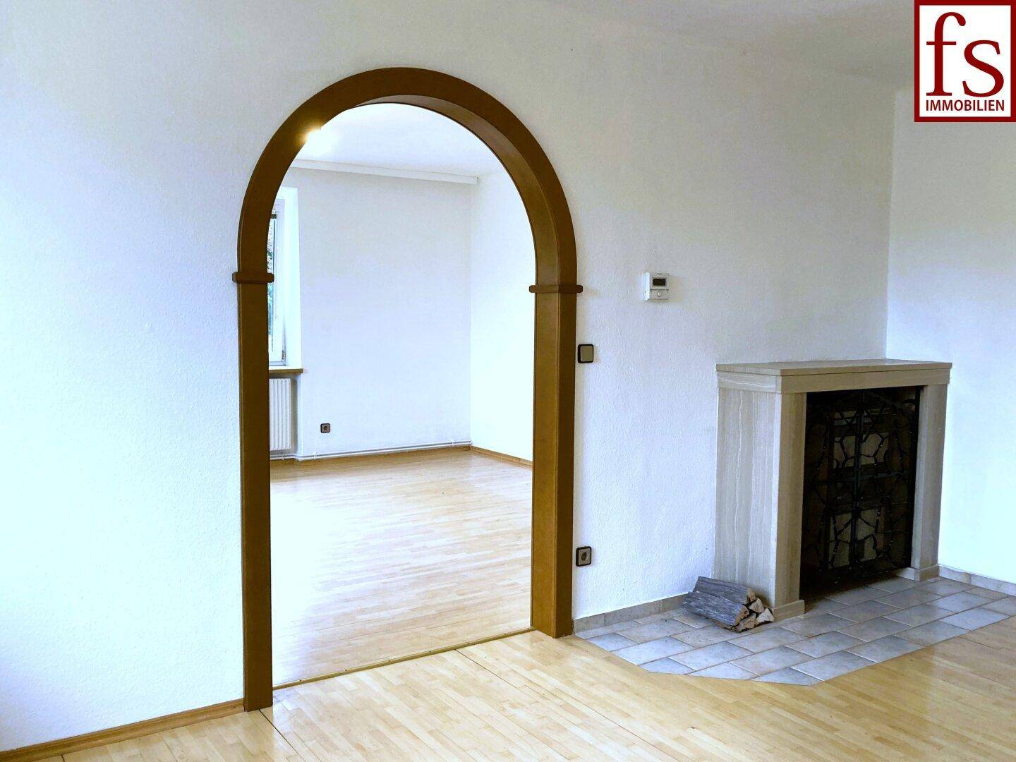 Wohnbereich mit Holzboden, dekorativem Kamin und einem Rundbogen, der zu einem weiteren Raum führt.