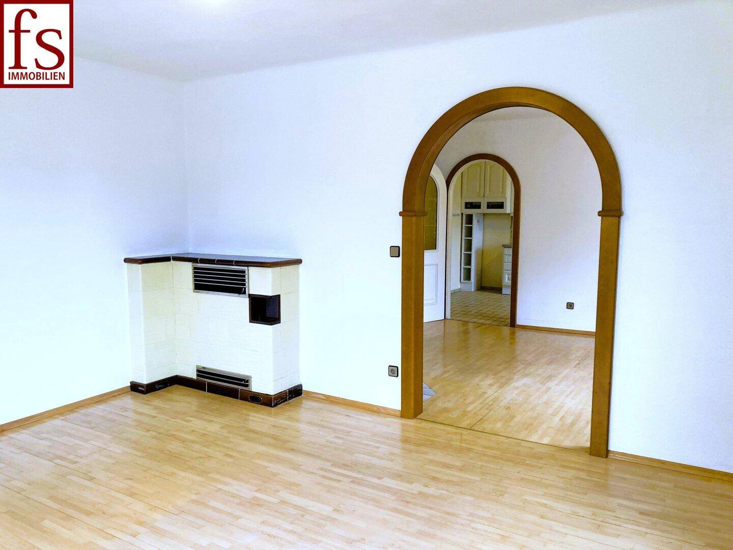 Geräumiger Wohnraum mit Parkettboden, traditionellem Kachelofen und Rundbogen zu einem weiteren Zimmer.