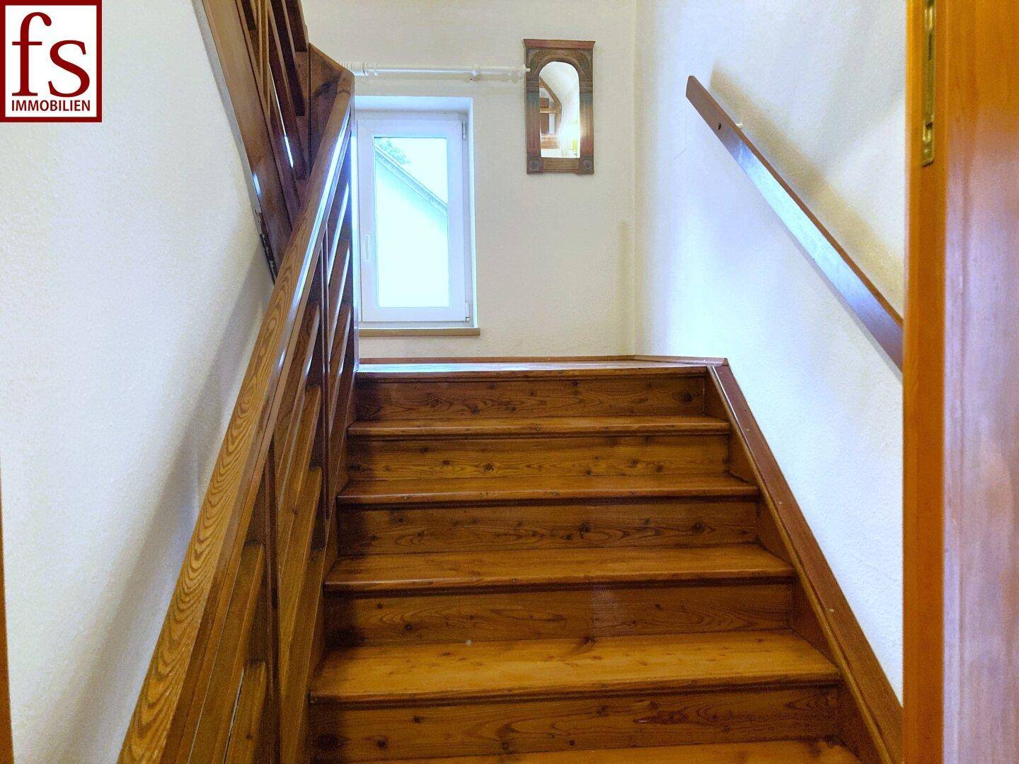 Holztreppe mit Geländer führt in das obere Stockwerk, beleuchtet durch ein Fenster.