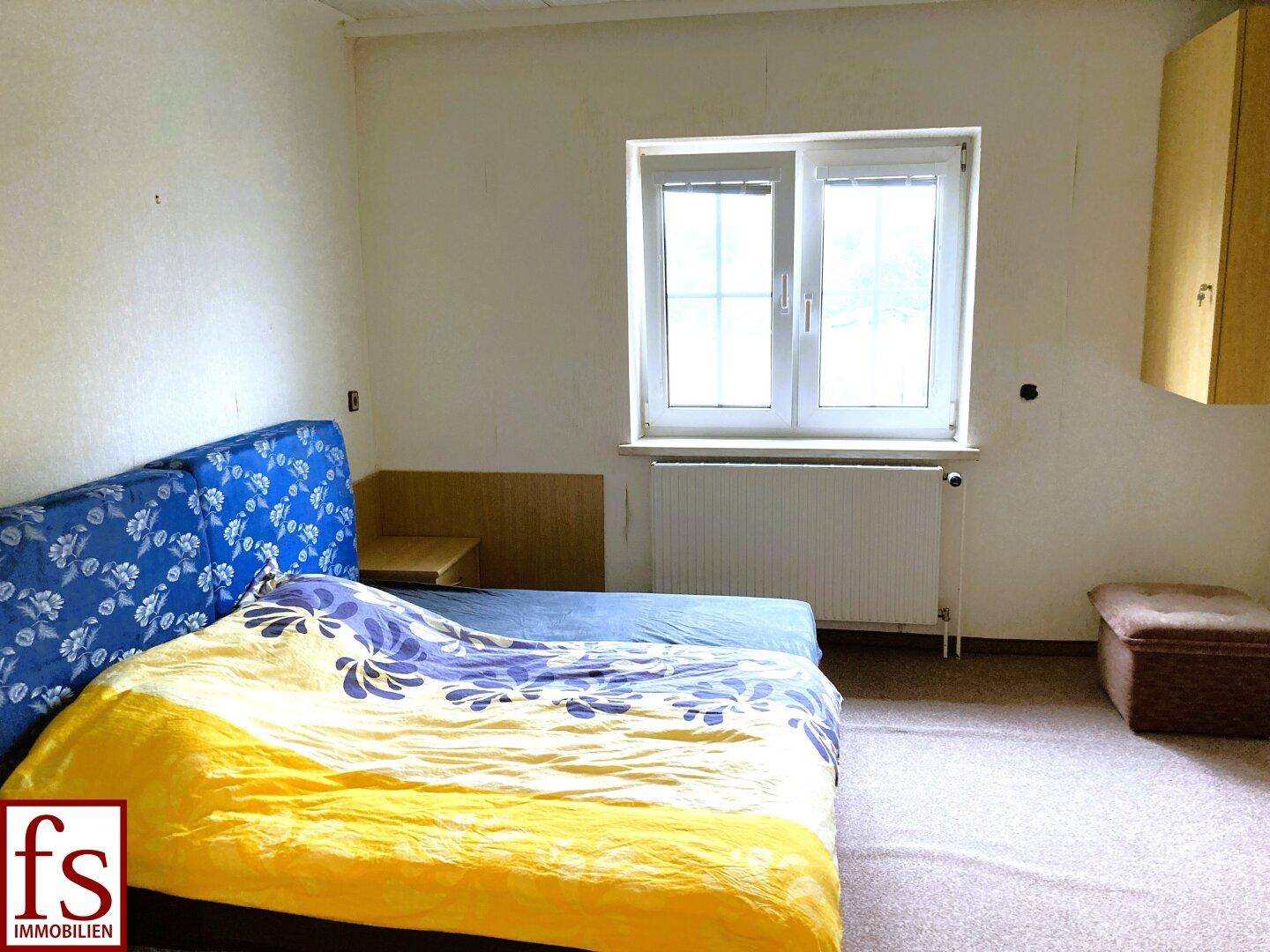 Schlafzimmer mit Teppichboden, Doppelbett und Fenster, das viel Tageslicht hereinlässt.