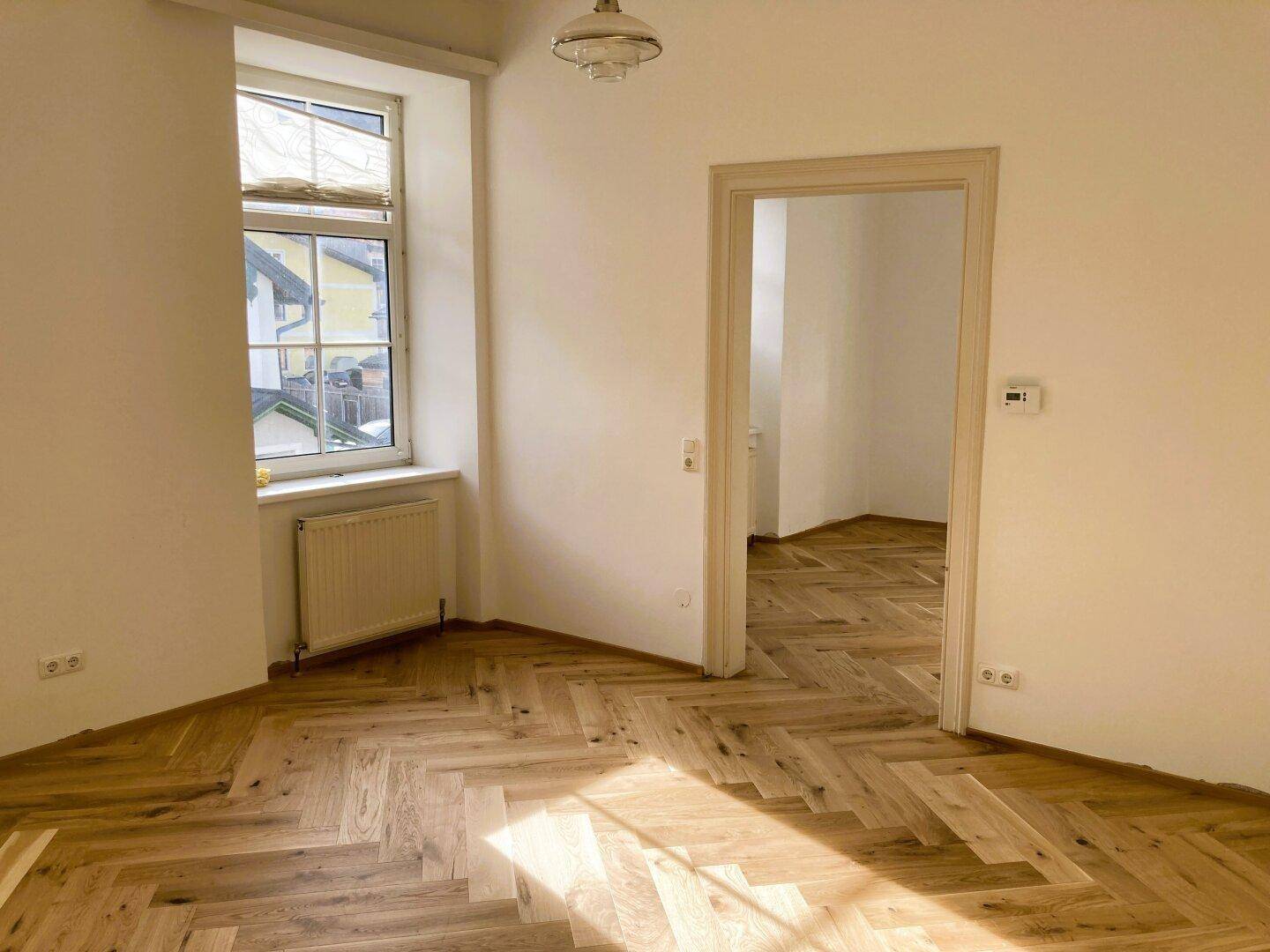 Helles Zimmer mit Fischgrätparkett, Heizkörper und Blick ins Grüne durch das Fenster.