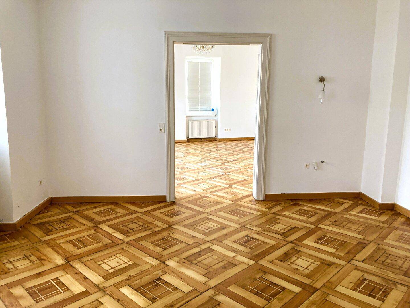 Geräumiges Zimmer mit Parkettboden und offenem Durchgang zu einem weiteren hellen Raum.