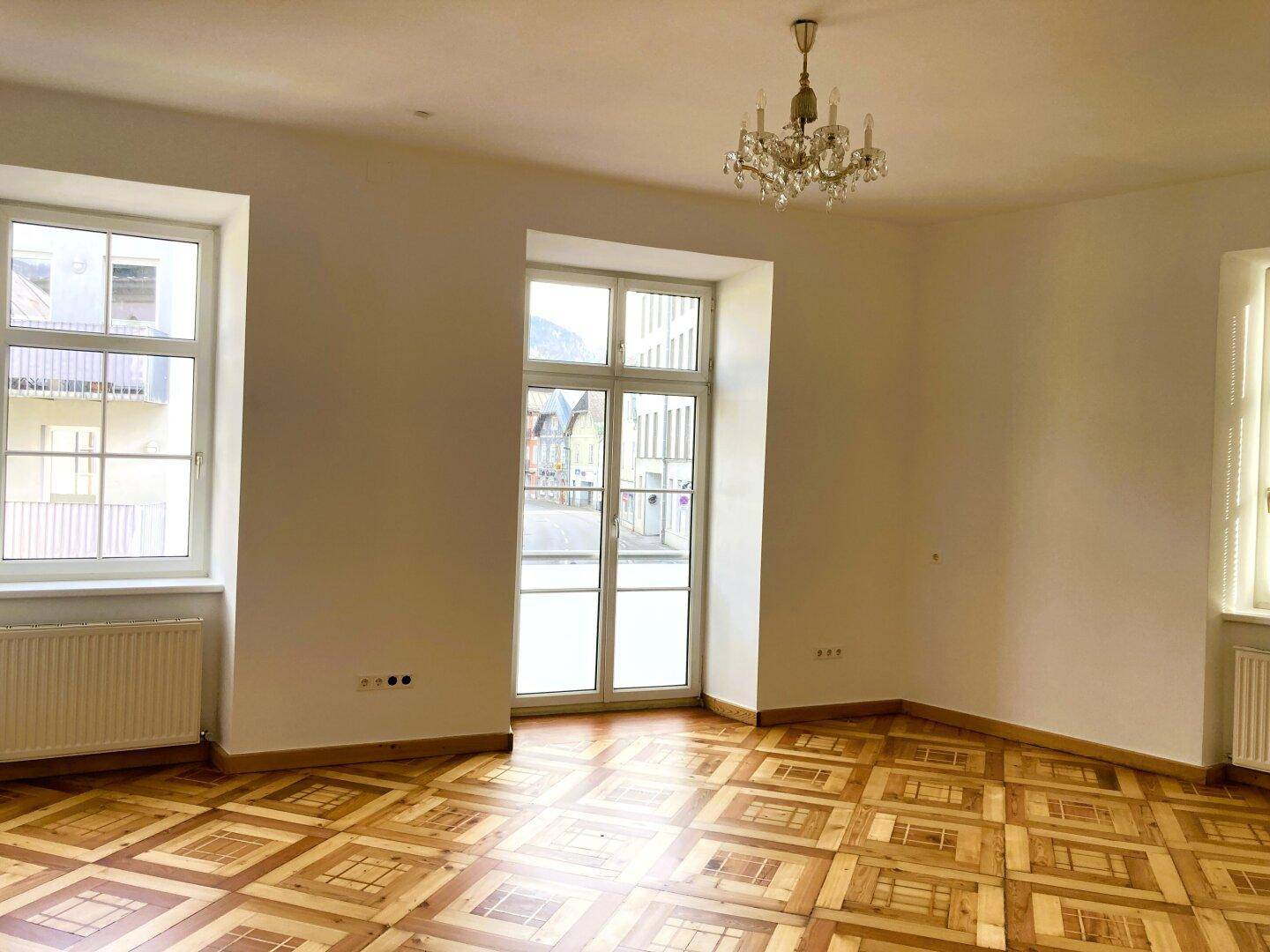 Heller Raum mit Parkettboden, Kronleuchter und Blick auf die Straße durch große Fenster.
