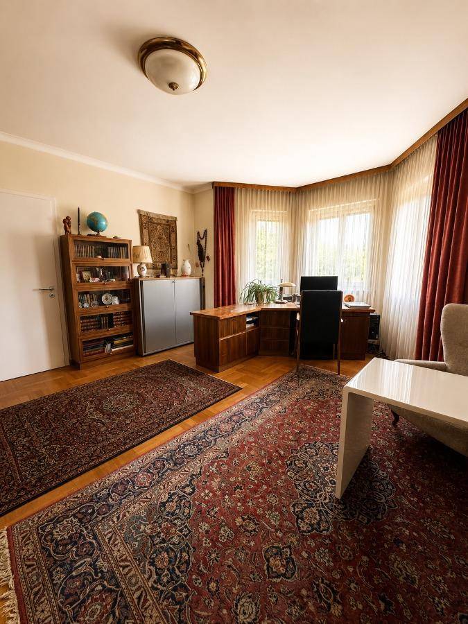 Geräumiges Büro mit Schreibtisch, Bücherregal und traditionellen Teppichen, hell durch Erkerfenster.