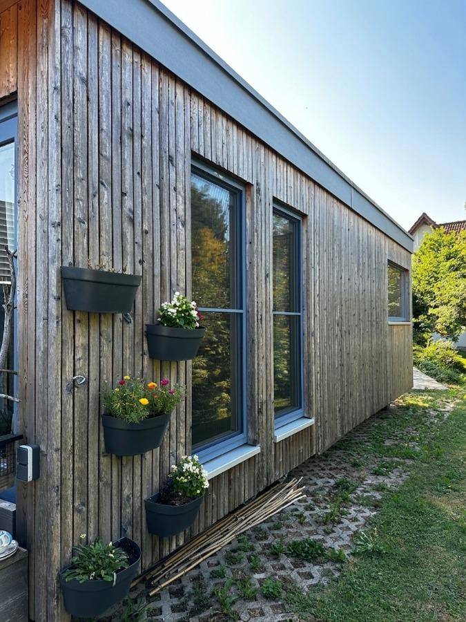 Seitenansicht des Holzhauses mit vertikalen Holzpaneelen und Blumenkästen an den Fenstern.