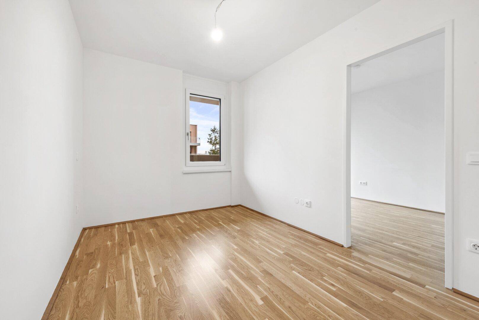 Helles, leeres Zimmer mit Parkettboden und einem Fenster, das Tageslicht hereinlässt.