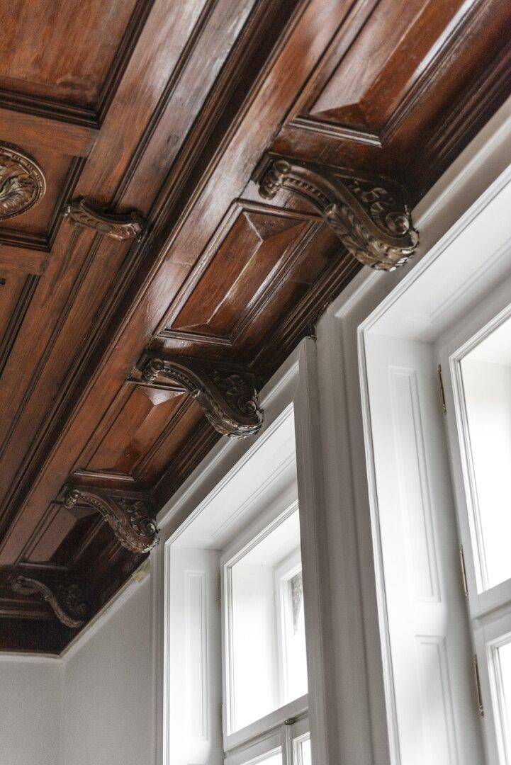 Detailaufnahme einer reich verzierten Holzkassettendecke und hohen Fenstern.