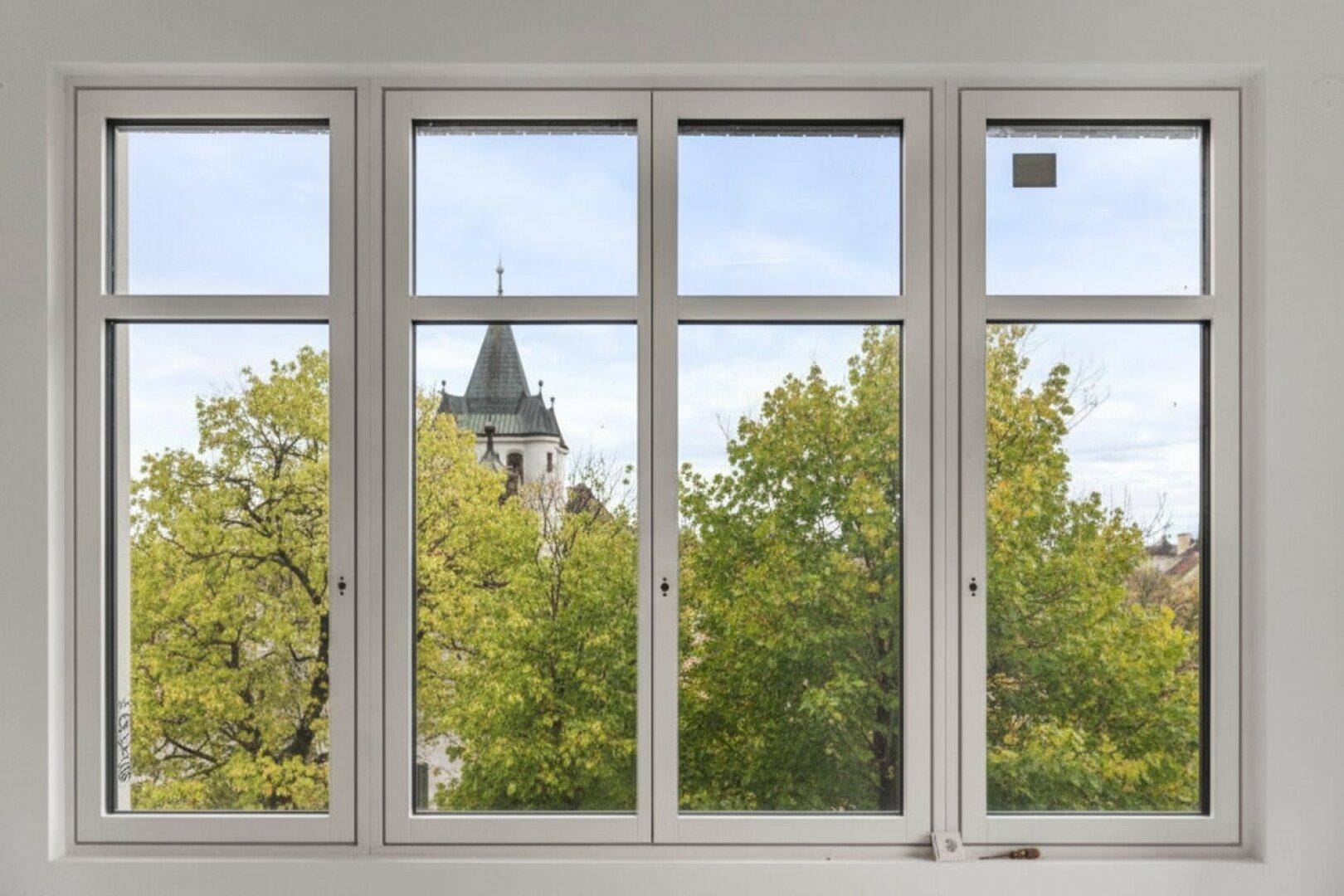 Fenster mit Blick auf grüne Bäume und einen historischen Turm im Hintergrund.