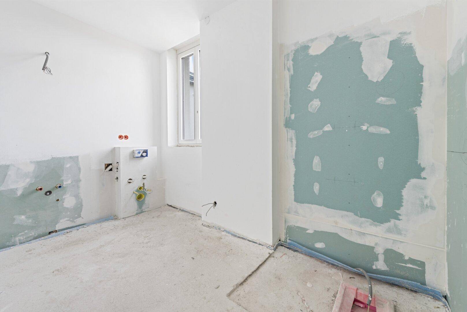 Badezimmer im Rohbau mit sichtbaren Installationen und einem Fenster, bereit für den finalen Ausbau.