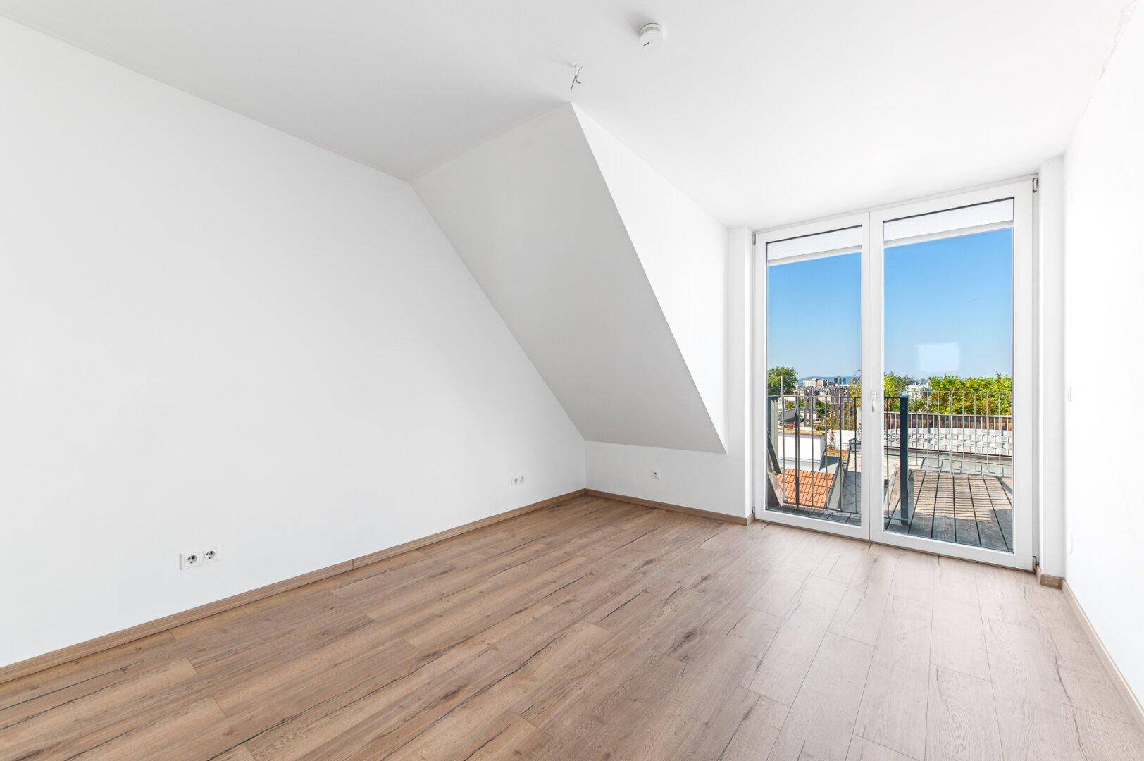 Heller Raum mit Holzboden, Dachschräge und Zugang zu einem Balkon mit Stadtblick.