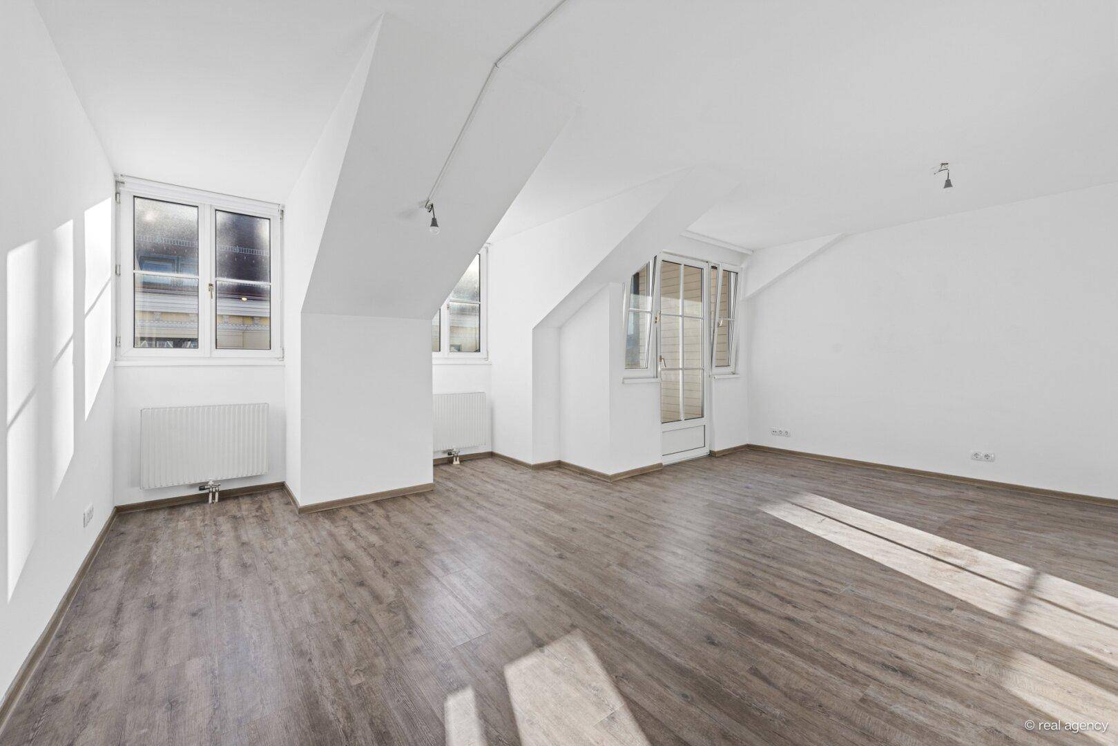 Lichtdurchfluteter Raum mit Holzboden, Dachschrägen und Zugang zu einem kleinen Balkon.