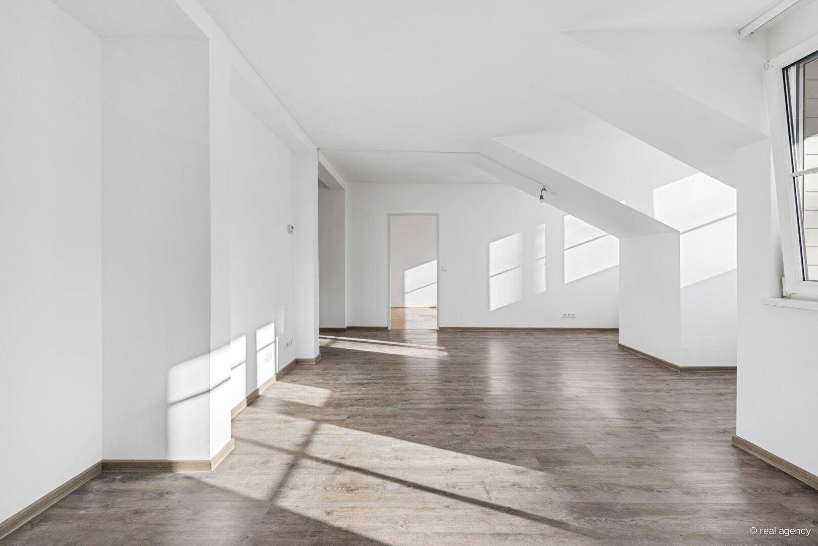 Heller, leerer Raum mit Holzboden, weißen Wänden und viel natürlichem Licht durch die Fenster.