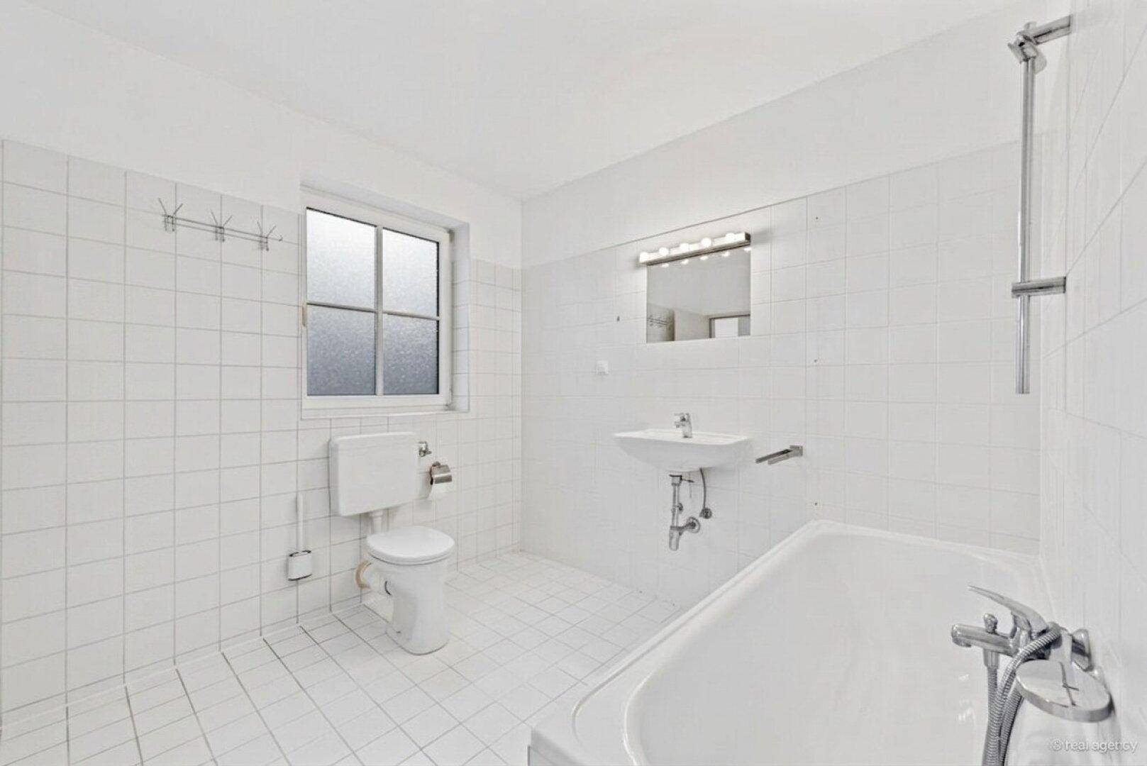 Helles Badezimmer mit weißen Fliesen, Badewanne, WC, Waschbecken und Fenster.