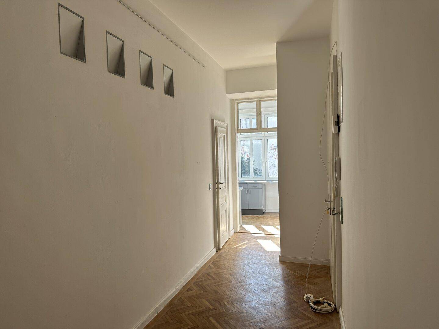 Langer Flur mit Fischgrätparkett, der zu einem hellen Raum mit Fenster und Tageslicht führt.