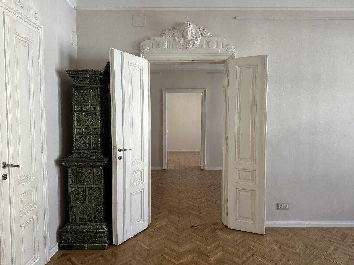 Blick durch eine offene, verzierte Tür in einen weiteren Raum mit Fischgrätparkett und Kachelofen.