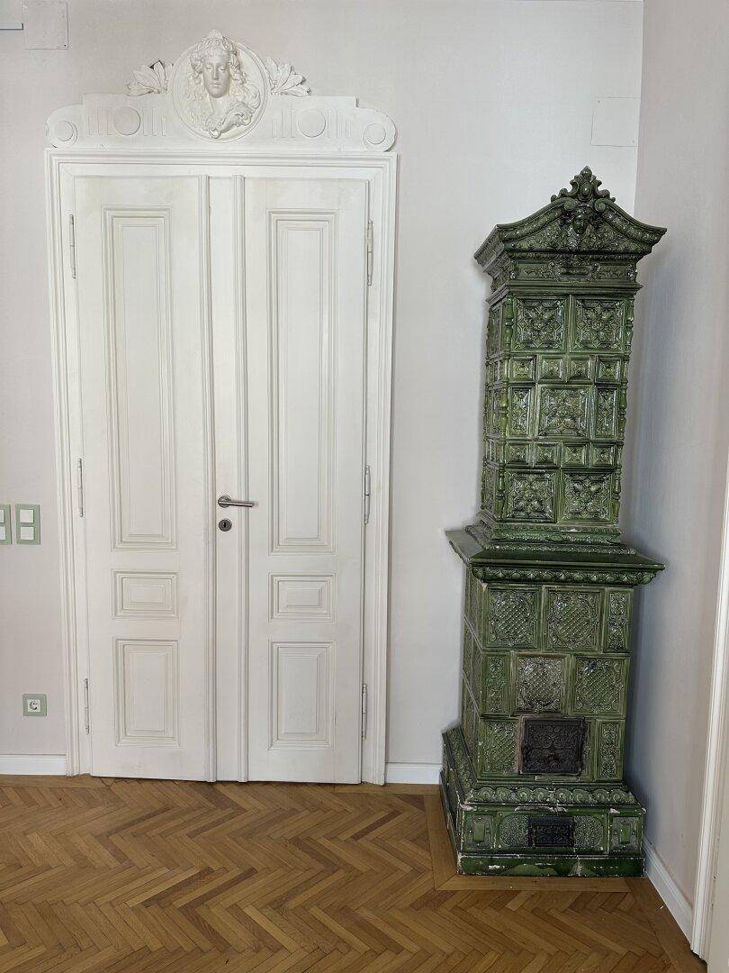Geräumiges Zimmer mit Fischgrätparkett, einer verzierten Tür und einem traditionellen grünen Kachelofen.