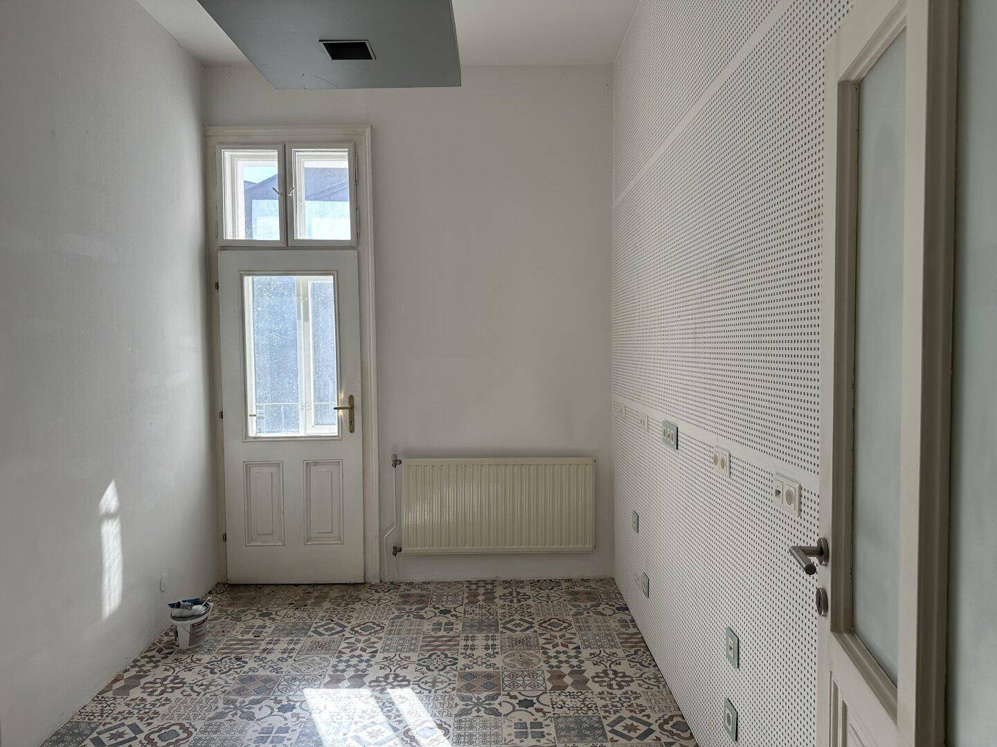 Kleiner Raum mit gemusterten Fliesen, Heizkörper und einer Tür mit Fenster, die Tageslicht spendet.