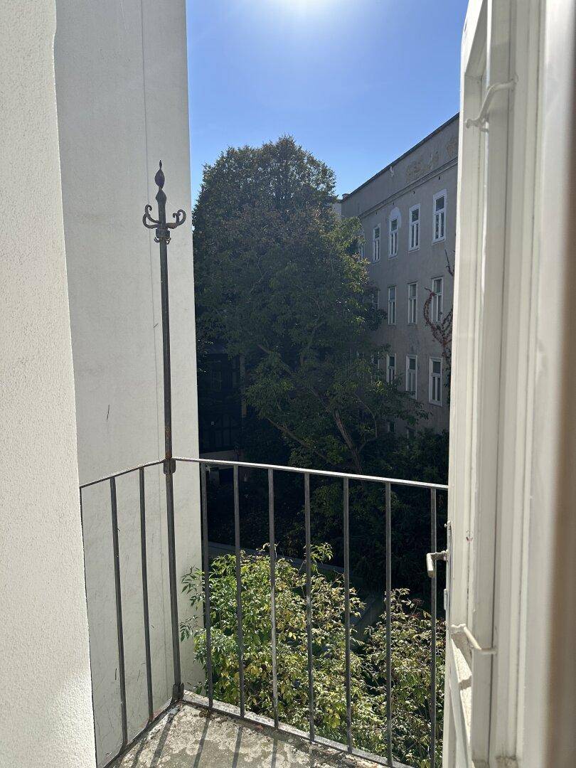 Kleiner Balkon mit Blick auf einen grünen Innenhof und alte Gebäude, umgeben von Bäumen.