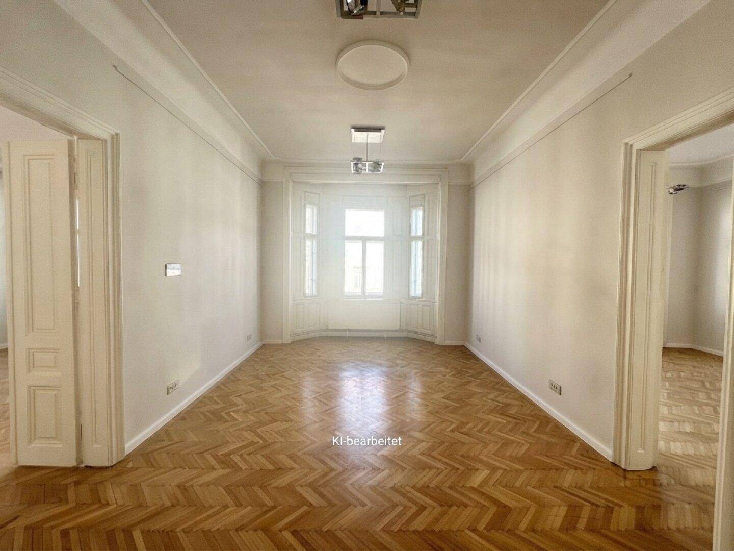 Heller, langer Raum mit Fischgrätparkett, hohen Decken und einem Erkerfenster, das viel Licht hereinlässt.