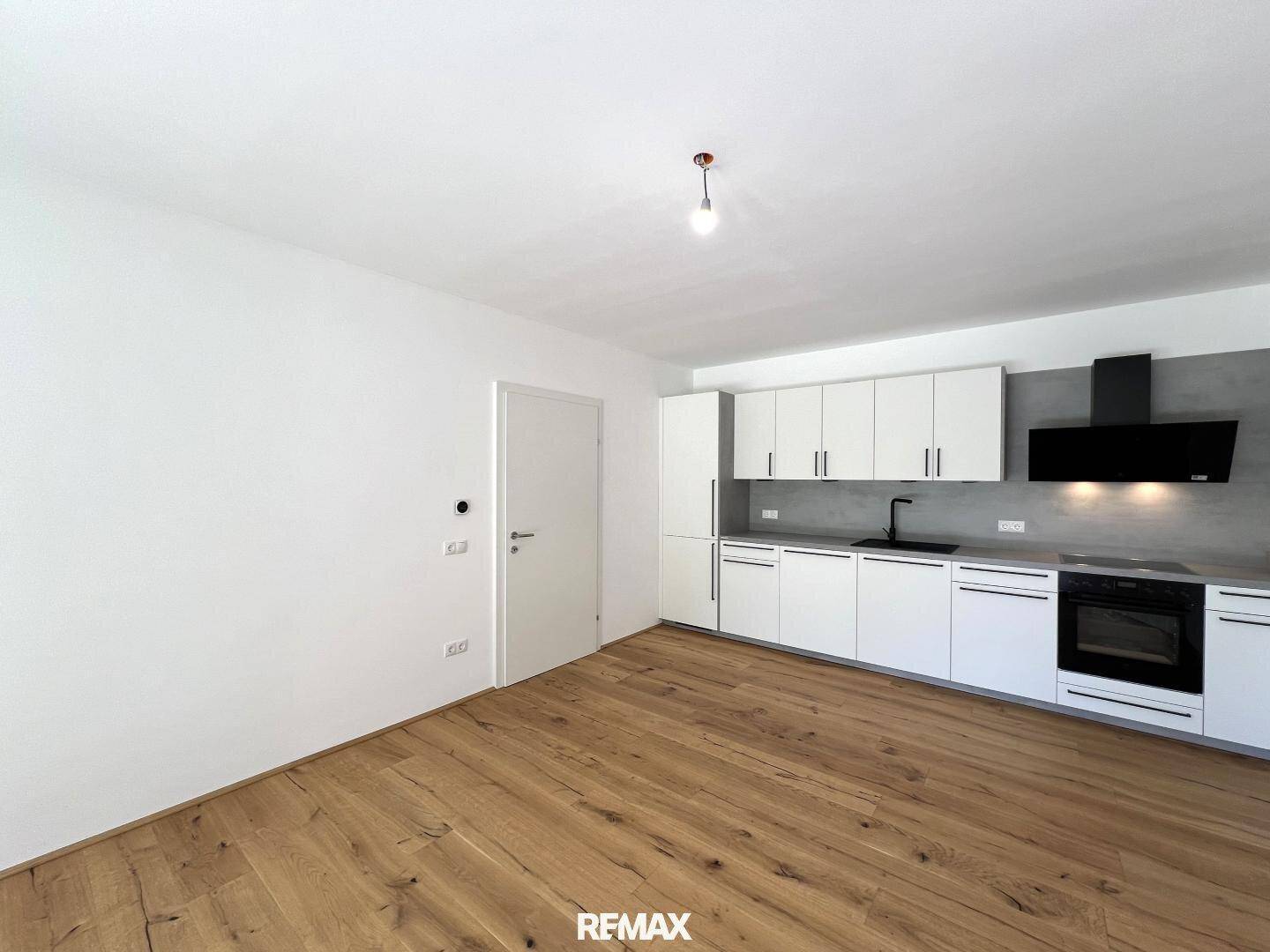 Moderne Einbauküche mit weißen Schränken, dunkler Arbeitsplatte und Holzboden in offenem Raum.